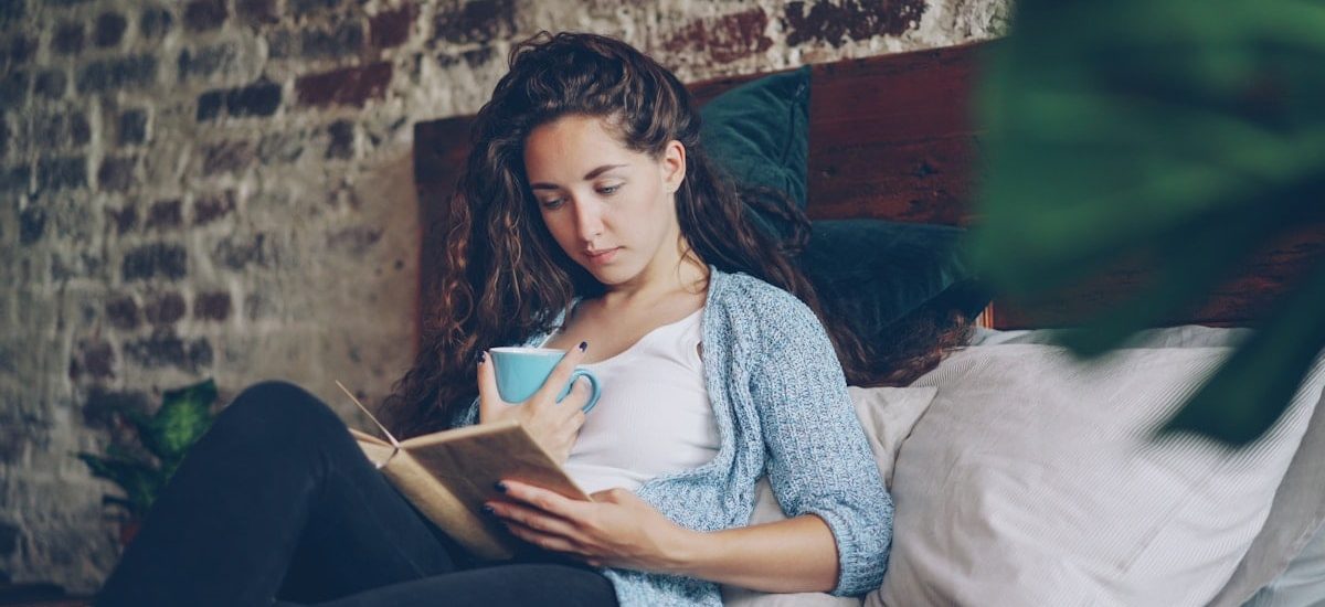 Cheerful girl is reading funny book in bedroom lying on comfy double bed, smiling and holding cup with drink. Hobby, happy people, beautiful interiors and literature concept.
