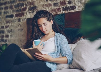 Cheerful girl is reading funny book in bedroom lying on comfy double bed, smiling and holding cup with drink. Hobby, happy people, beautiful interiors and literature concept.